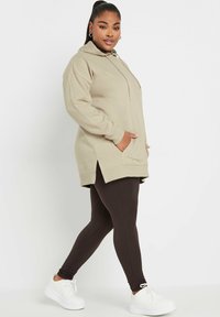 Hoodie beige oversize con tasca frontale, fessure laterali e cordini, abbinato a leggings marroni a costine e sneakers bianche.