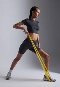 Black cropped athletic shirt and shorts, gripping yellow resistance bands. Gray background, person in action, wearing gray and yellow sneakers.