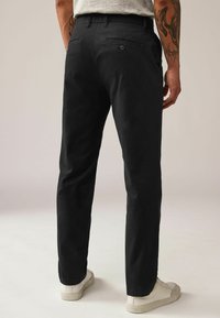 Pantaloni negri cu un design drept, având buzunare laterale cu închidere cu nasturi. Purtați cu adidași albi pe un fundal neutru.