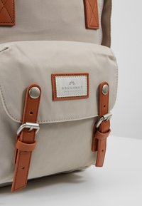 Sac à dos en toile beige avec des accents en cuir, fermeture à bouton et un patch de marque cousu. Comprend des bretelles en cuir ajustables et des accessoires métalliques.