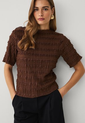 Kvinde med langt bølget hår iført en struktureret brun kortærmet top og sorte bukser, stående med hænderne i lommerne mod en hvid væg.