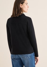 Camisa negra de manga larga con una textura suave, con cuello y detalles de costura en la parte posterior, combinada con jeans azules.