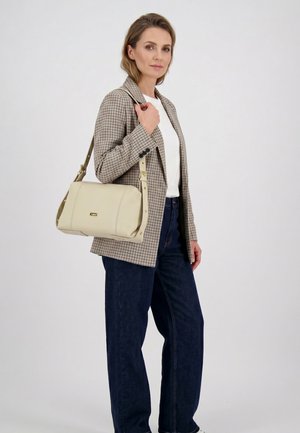 Kvinde i ternet blazer, hvid skjorte og mørke jeans med en beige skuldertaske, stående foran en ensfarvet lys baggrund.