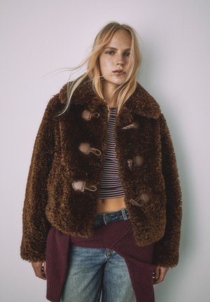 Hnedá bunda z umelého kožúšku so širokým golierom a zapínaním na gombíky, nosená cez pásikavý krátky top, skombinovaná s denimovými nohavicami.