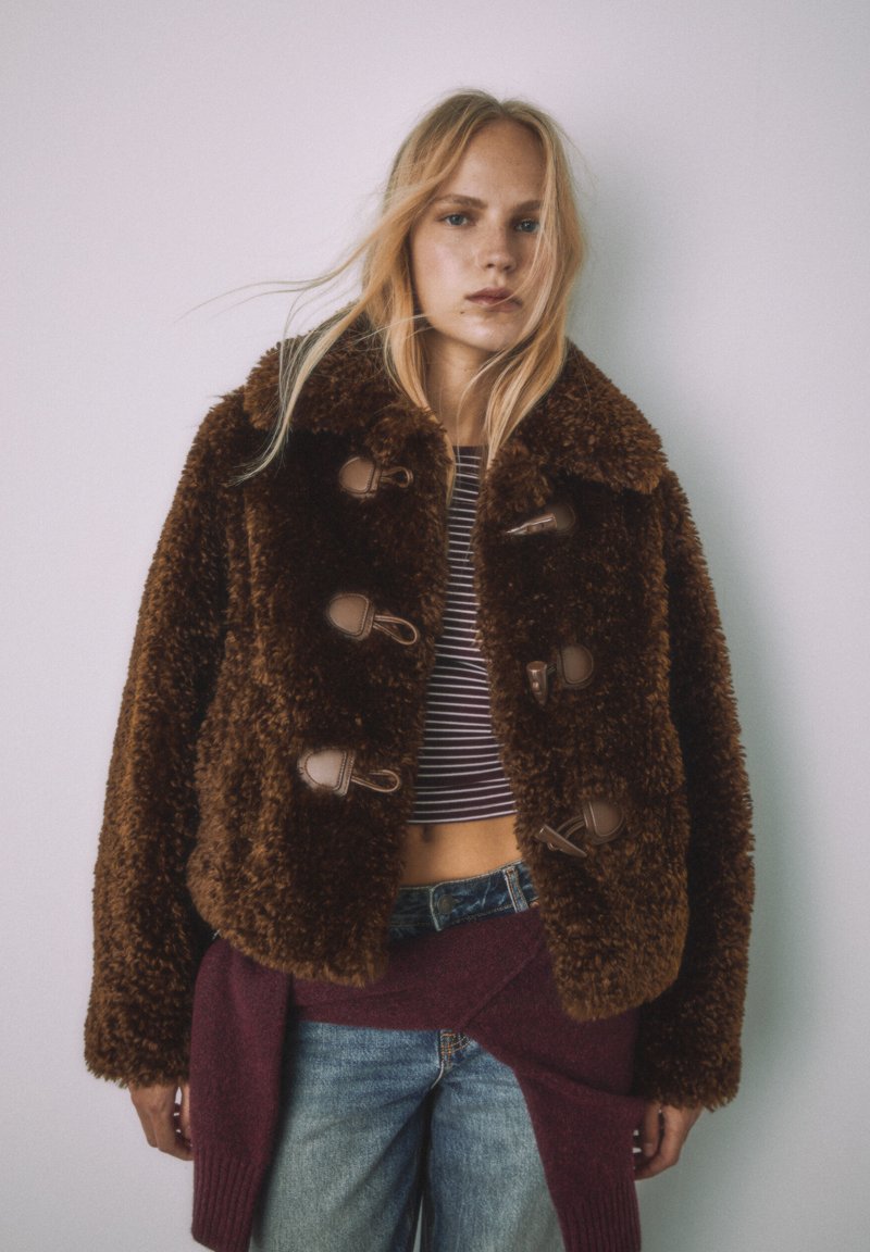 Brun jacka i konstpäls med bred krage och knäppning med knappar, bärs över en randig crop top, kombinerad med jeans.