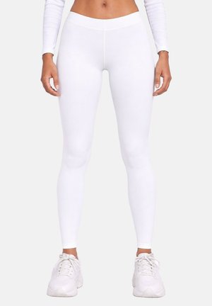 Femme portant des leggings blancs taille haute et des baskets blanches, debout les mains détendues le long du corps sur un fond uni.