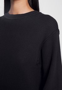 Maglione nero lavorato a maglia con una texture a coste, scollatura rotonda e spalle scese. Presenta una piccola apertura laterale per un tocco in più.