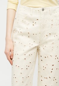 Κοντινό πλάνο λευκών παντελονιών ψηλόμεσων με κεντήματα ανθικών μοτίβων, φορεμένα με ανοιχτοκίτρινη ριμπ κορυφή.