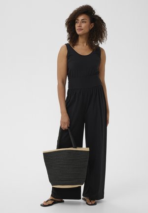 Femme aux cheveux bouclés portant une combinaison sans manches noire, tenant un grand sac fourre-tout tissé noir et beige, avec des sandales noires.