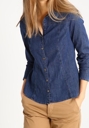 Vrouw draagt een nauwsluitend donkerblauw denim overhemd met knopen en driekwartmouwen, met beige broek, handen in de zakken.
