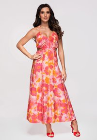 Femme portant une robe maxi sans manches rose et orange à fleurs, avec des découpes à la taille et des sandales rouges à talons hauts, debout avec une main sur la hanche.