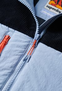 Giacca azzurra chiara con colletto rigato nero testurizzato, doppie zip arancioni e corpo in tessuto liscio a contrasto con cuciture visibili.