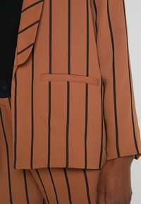 Braun gestreifter Blazer mit schwarzen vertikalen Linien, ausgestattet mit einem Revers, einer einzelnen Brusttasche und glatter Stofftextur.