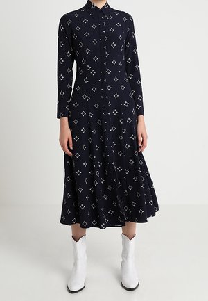 Granatowa sukienka midi z długim rękawem w biały wzór diamentów, z zapięciem na guziki z przodu, noszona z białymi, sięgającymi połowy łydki, spiczastymi kowbojkami.