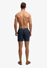 Homme debout, pieds nus, de dos, portant un short de bain bleu marine et des sandales marron, sur fond blanc uni.