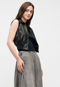 Νεαρή γυναίκα με ίσια καστανά μαλλιά φοράει αμάνικο μαύρο δερμάτινο γιλέκο, μαύρο τοπ και γκρι υφασμάτινη φούστα με ψηλή μέση, στέκεται πλαγίως.