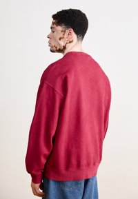 Felpa rossa realizzata in morbido cotone, con una vestibilità ampia, spalle dropped e polsini a costine. Indossata con jeans di denim blu.