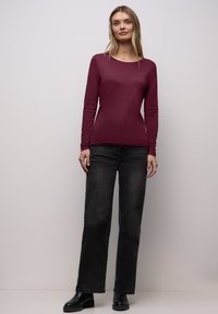 Bordeauxrotes Langarmoberteil aus weichem Material, kombiniert mit schwarzen Jeans mit weitem Bein und schwarzen Stiefeln. Einfacher Rundhalsausschnitt, figurbetontes Design.