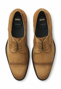 Paar beige Wildleder-Schnürschuhe mit Brogue-Details an den Zehenkappen und "BOSS Crafted in Italy" auf den schwarzen Innensohlen gedruckt.