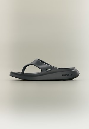 Sandale tong slip-on gris foncé Lacoste avec une semelle épaisse et des trous de ventilation latéraux, présentée de profil sur un fond beige.