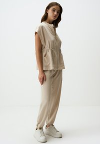 Completo beige in due pezzi composto da un top corto con zip e maniche corte, con vita a cordoncino e due tasche frontali, abbinato a pantaloni jogger affusolati coordinati.