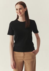 T-shirt noir à manches courtes, coupe ajustée, en tissu lisse, associé à un pantalon marron clair, présenté sur un fond neutre.