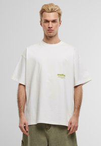 White oversized t-shirt made of cotton, features short sleeves, a crew neckline, and a small green logo on the left chest area.