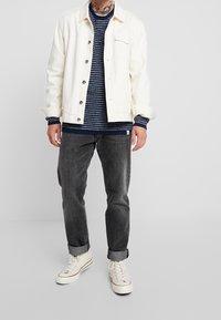 Giacca di jeans bianca, maglione navy testurizzato con strisce, jeans grigio scuro e sneaker bianche, con risvolti e dettagli a bottone.