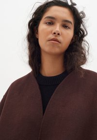 Rudas vilnonis paltas su lygiu paviršiumi, turintis platų apykaklę ir atvirą priekį. Dėvimas ant juodo megztinio su grioveliu apykaklėje.