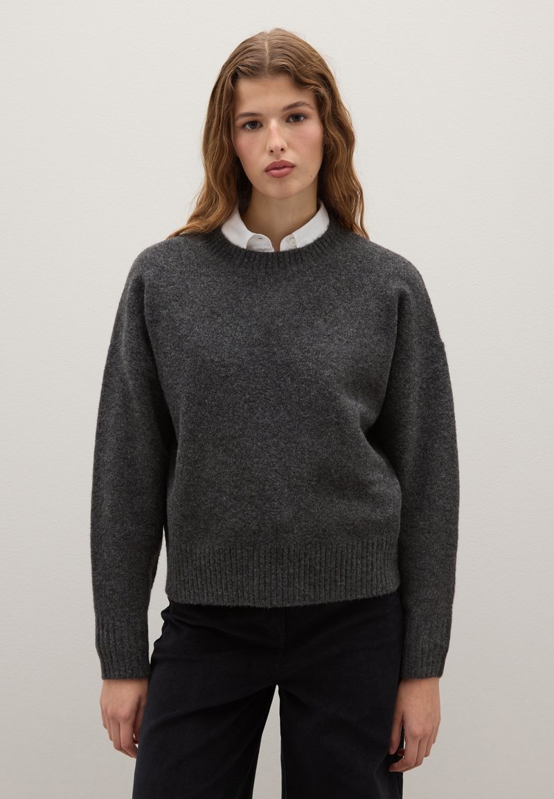 Dunkelgrauer Wollpullover mit rundem Halsausschnitt, geripptem Saum und lockerer Passform. Kombiniert mit einem weißen Hemd darunter.