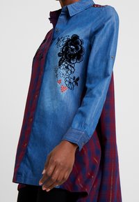 Donna che indossa una camicia in chambray blu a maniche lunghe con stampa floreale nera e cuori rossi, abbinata a un pannello posteriore a quadri rossi e blu navy.
