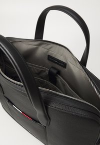 Schwarze Lederhandtasche mit einem strukturierten Design, Reißverschluss und zwei Griffen. Das Innere ist mit einem grauen Futter und einem kleinen Markenschild ausgestattet.