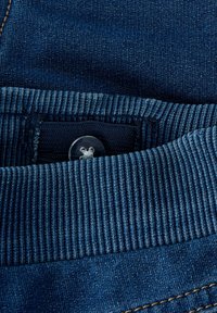 Pantalons en denim bleu avec une taille côtelée, une fermeture à bouton argenté et des détails de couture visibles. Le tissu présente un tissage texturé.