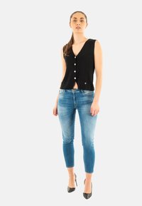 Gilet lavorato a maglia nera con scollatura a V, caratterizzato da cinque bottoni bianchi e un piccolo dettaglio a forma di cuore. Abbinato a jeans skinny blu chiaro e contrasti neri.