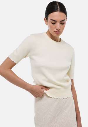 Pull crème à manches courtes avec un col rond, texture côtelée et bords festonnés. Associé à une jupe beige détaillée.