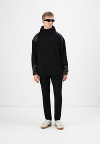 Μαύρη φαρδιά hoodie με υφή στους μαύρους ώμους, συνδυασμένη με στενά μαύρα παντελόνια και λευκά αθλητικά παπούτσια. Το μοντέλο φοράει σκούρα γυαλιά ηλίου.