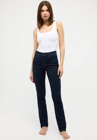 Mørkeblå høytplastiske jeans med smal passform, med frontlommer og synlige sømmer; parret med en tettsittende hvit tanktopp.