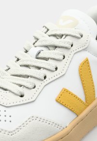Sneaker in pelle bianca con logo accentato giallo testurizzato, suola beige e lacci grigi. Presenta perforazioni sui lati e punta arrotondata.