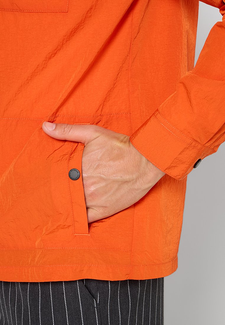 Hand in der Tasche einer orangefarbenen Langarmjacke mit Druckknöpfen, kombiniert mit schwarzen gestreiften Hosen.