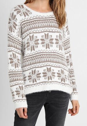 Femme portant un pull en maille blanc et beige avec un motif flocon de neige et un jean noir, debout devant un fond clair uni.