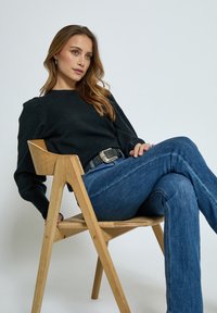 Black knit sweater with puff sleeves, worn with faded blue flared jeans. Sitting on a light wood chair with a natural finish. Silver-buckled belt.
