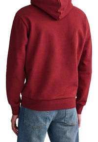 Persoon gekleed in een effen rode hoodie en blauwe spijkerbroek, gezien van achteren.