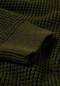 Tissu en maille vert foncé, présentant un motif texturé avec des poignets côtelés. Matière douce et épaisse offrant une apparence confortable.