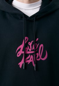 Felpa con cappuccio nera con un logo scritto in rosa ben visibile sul davanti. Realizzata in tessuto strutturato con coulisse regolabili.