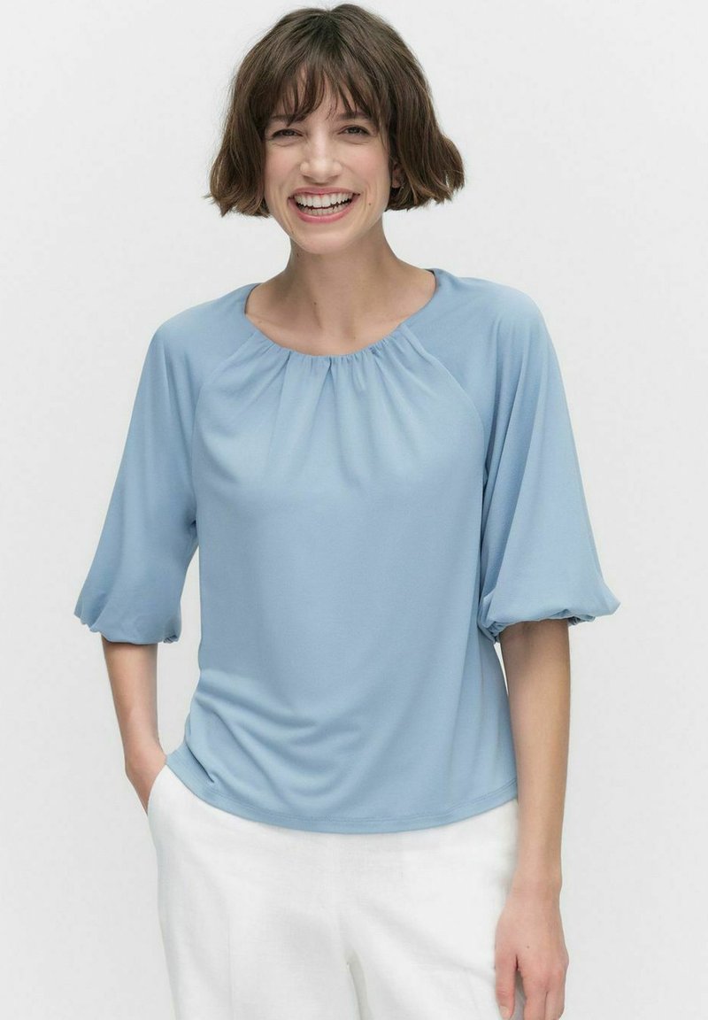 Lyseblå bluse med rund halsudskæring, pufærmer og løs pasform. Fremstillet af blødt stof med en glat tekstur. Kombineret med hvide bukser.