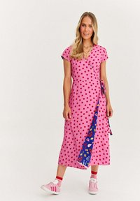 Růžové zavinovací šaty s červenými puntíky, krátké rukávy a kontrastní modrý vzorovaný doplněk. Vydáno s růžovými teniskami a ponožkami s červenými pruhy.