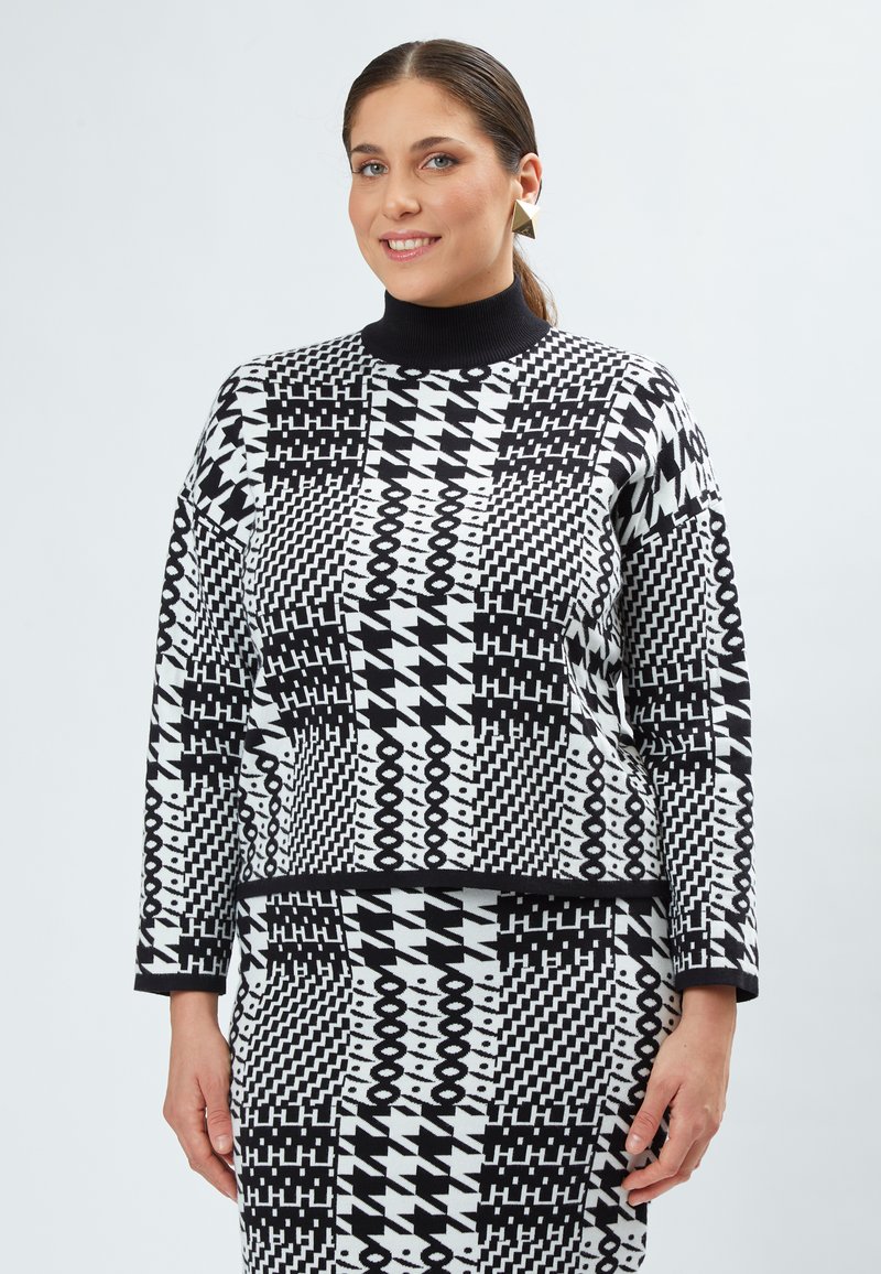 Sort-hvid mønstrede sweater med høj krave, der præsenterer geometriske former og varierende teksturer. Model poserer mod en lys baggrund.