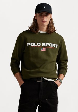 Hanorac de culoare verde măsliniu cu textul "POLO SPORT" în alb și roșu, având un detaliu cu steagul american. O șapcă neagră și pantaloni închisi la culoare completează aspectul.