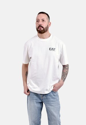 Uomo con barba e tatuaggi che indossa una t-shirt bianca EA7 Emporio Armani e jeans azzurri, in piedi con una mano in tasca.