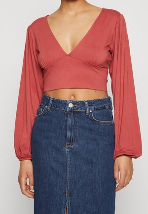 Vrouw draagt een roestkleurig cropped top met lange mouwen en een diepe V-hals, gecombineerd met een donkerblauwe denim rok met hoge taille en een voorwaartse split.